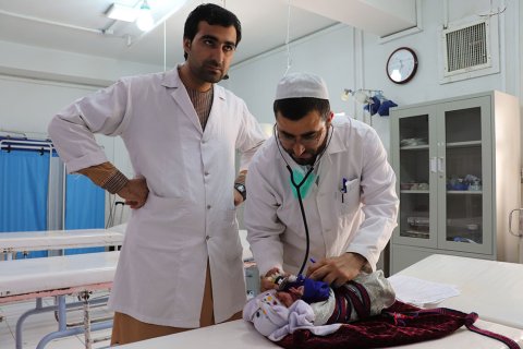 Två män iklädda vita sjukhusrockar står vid en brits på Boost sjukhus, Lashkar Gah, Afghanistan. På britsen ligger en liten bebis.