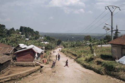 Personer går längs en slutande grusväg. På sidorna syns enklare hus i trä. 