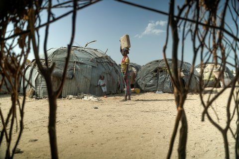 Framför små hyddor står en kvinna hållandes en vattendunk på huvudet.