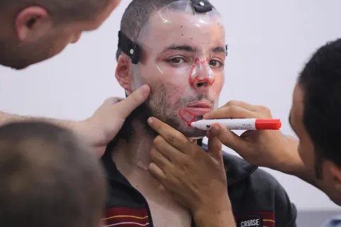 En man har en plastmask för ansiktet. En annan personer målar på masken med en röd penna.