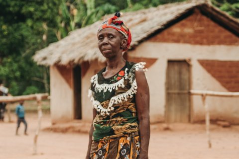 En medelålderskvina står med en neutral blick tittandes mot kameran, i bakgrunden syns ett tegelhus i ett stepplandskap.