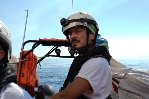 En man med flytväst och hjälm kollar mot kameran medan han står i en båt med öppet hav bakom ryggen