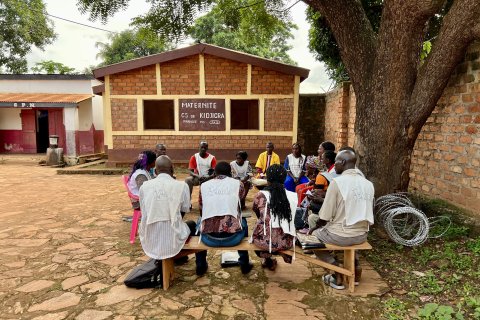 en grupp vårdpersonal på en hälsocentral sitter på träbänkar under ett träd utanför byggnaden