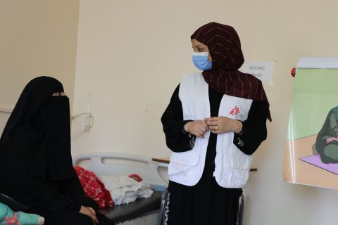 En kvinna med Läkare Utan Gränser-väst och munskydd står vid en säng där en annan kvinna sitter. Till höger ser man en illustration av en familj som sitter på en filt och äter. 