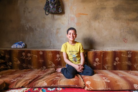 En leende pojke i 10-årsåldern med gul t-shirt sitter på en madrass på golvet i ett kalt rum