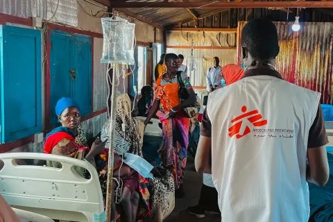 Ryggtavla av MSF-anställd omringad av patienter på en klinik.