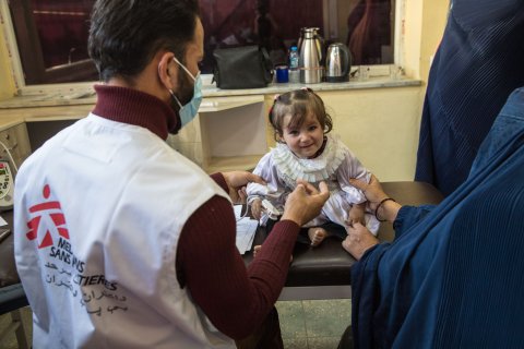 en liten flicka sitter på en brist på ett sjukhus, en manlig sjuksköterska och flickans mamma håller i henne