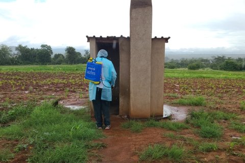 En person med skyddskläder och en container på ryggen står vid en liten betongbyggnad.