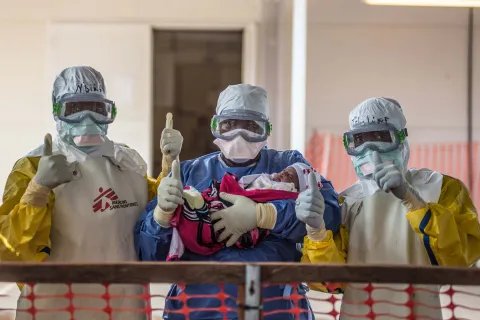 Tre vårdpersonal i skyddsdräkter från vårt ett av våra ebolacentrum gör tummen upp med ett spädbarn i famnen