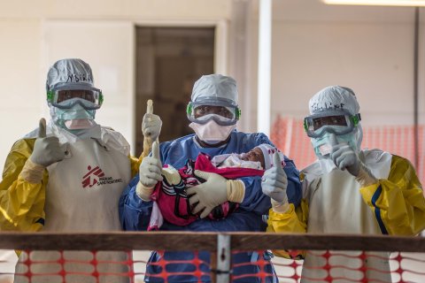 Tre vårdpersonal i skyddsdräkter från vårt ett av våra ebolacentrum gör tummen upp med ett spädbarn i famnen