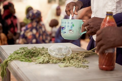 Ett bord med en grön växt/krydda på och händer som vispar i en kopp. Ur fokus i bakgrunden synds människor.