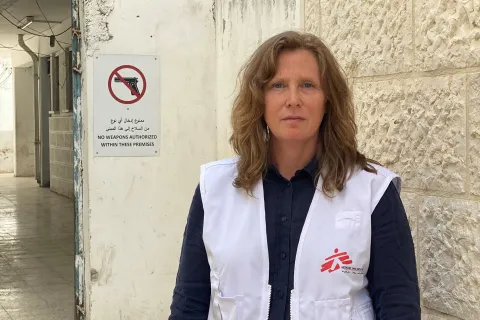 Kvinna tittar in i kameran, iklädd i Läkare Utan Gränser väst, i bakgrunden syns en skylt med vapen och ett sträck över.
