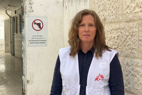 Kvinna tittar in i kameran, iklädd i Läkare Utan Gränser väst, i bakgrunden syns en skylt med vapen och ett sträck över.