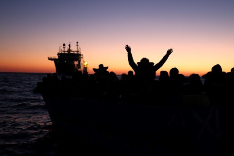 Skuggor av människor syns i skymningen och en båt syns i bakgrunden.