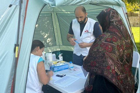 Två personer i Läkare Utan Gränser-västar och en kvinna vid ett tält med ett bord i.