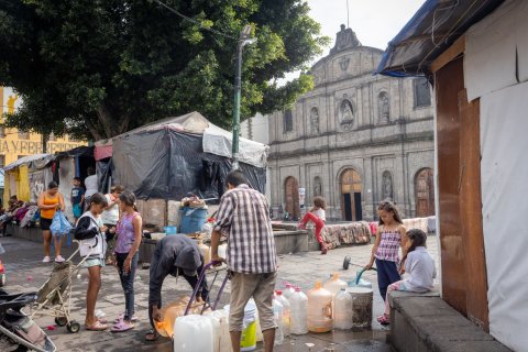 Människor hämtar vatten i stora dunkar på ett torg.