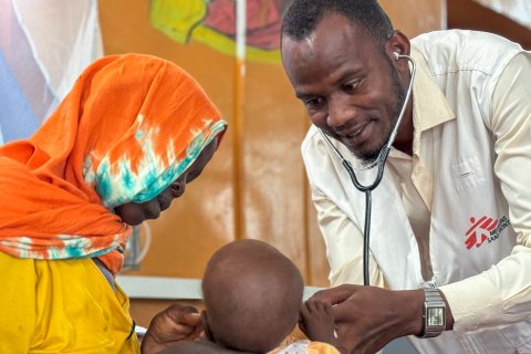 En man med stetoskop och Läkare Utan Gränser-väst undersöker ett barn. En kvinna är med barnet.