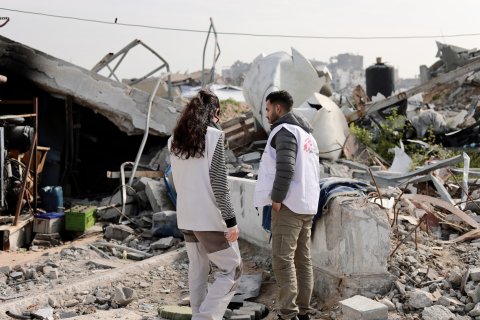 En man och en kvinna från Läkare Utan Gränser står framför rasmassorna av en byggnad som sprängts.