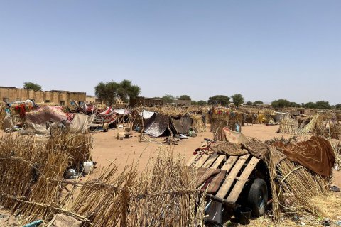 Provisoriska skydd gjorda av gräs, pinnar och tyg i ett läger i Sudan, med människor som rör sig mellan husen under en klar himmel.