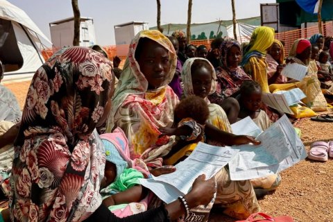 Kvinnor och barn sitter i skugga utanför klinik