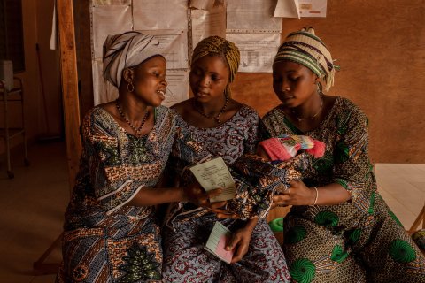 Tre kvinnor i mönstrade klänningar sitter på en bänk. En av kvinnorna räcker över ett spädbarn till en av de andra. 