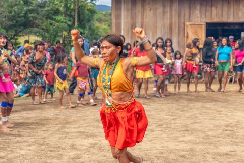 En kvinna i färgglada kläder har armarna upp i luften som att hon hoppar eller dansar. I bakgrunden står människor uppradade.