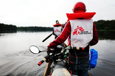 Två personer transporterar motorcyklar i en kanot över floden.