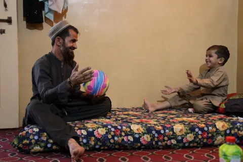 En man och ett barn sitter på en madrass. Mannen håller i en boll och barnet ler.