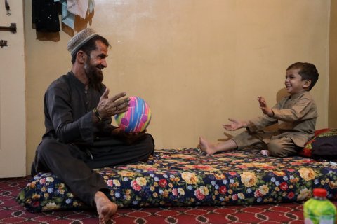 En man och ett barn sitter på en madrass. Mannen håller i en boll och barnet ler.