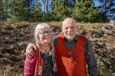 En man och en kvinna står utomhus i solen, mannen håller armen om kvinnans axlar och de tittar in i kameran.