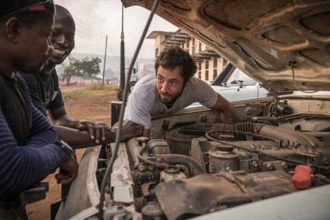 En man mekar med en bilmotor samtidigt som han pratar med två skrattande kollegor.