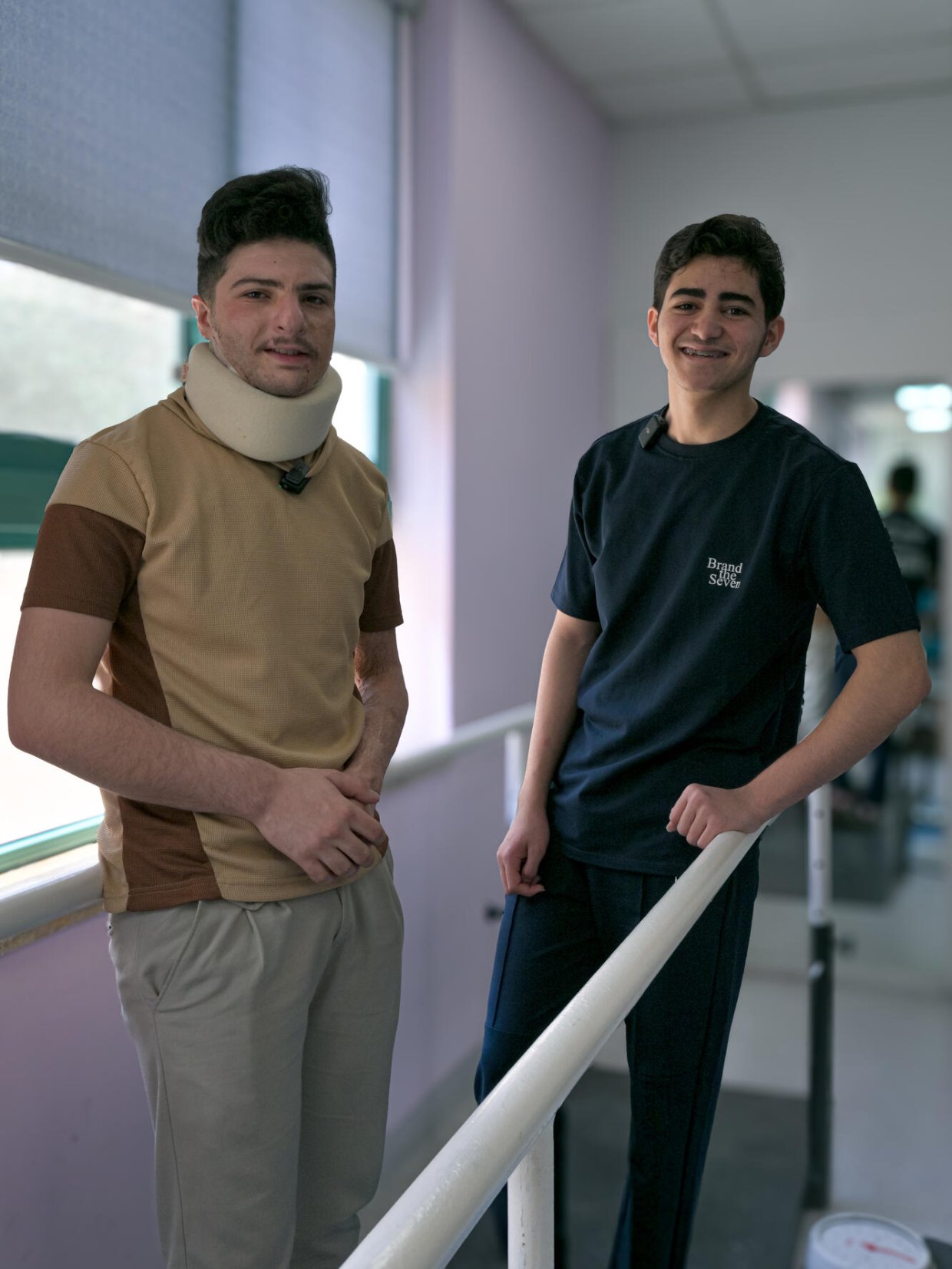 Två unga män står vid ett räcke inomhus. Den vänstra har en nackkrage runt halsen.