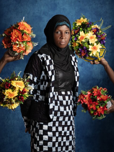 Kvinna står framför himmelsk bakgrund, omringas av blombuketter och tittar in i kameran.