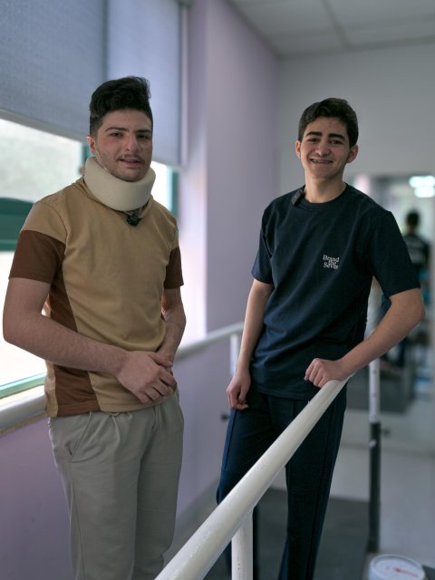 Två unga män står vid ett räcke inomhus. Den vänstra har en nackkrage runt halsen.