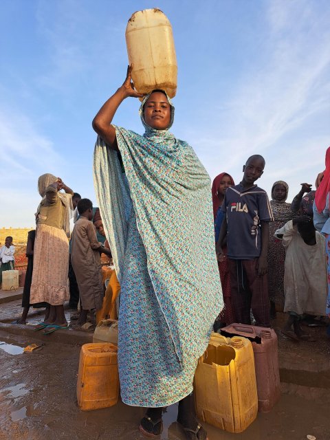 En kvinna med en vattendunk på huvudet. Barn i bakgrunden. Bilden tagen nedifrån.
