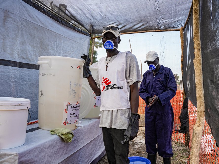En man med munskydd och Läkare Utan Gränser-väst står vid vita plasthinkar.