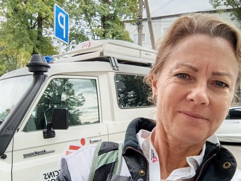 porträttbild på kvinna i Läkare Utan Gränser-väst framför en vit bil med Läkare Utan Gränser-logga.