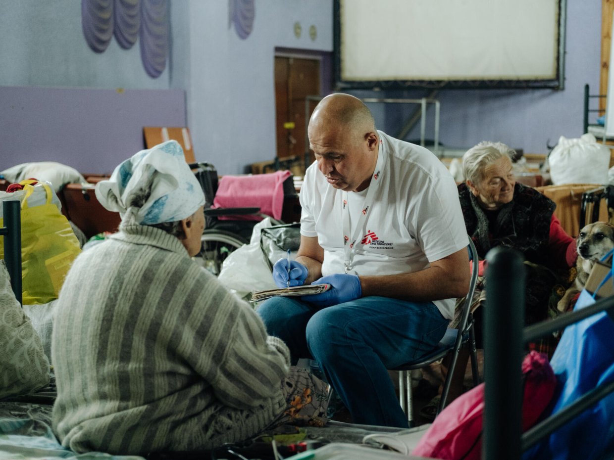 En man med Läkare Utan Gränser-väst sitter på en stol framför en kvinna som man ser ryggen på. I knät har mannen ett anteckningsblock som han skriver i. 
