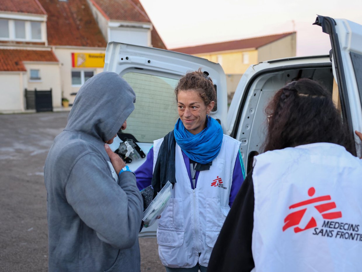 En kvinna i Läkare Utan Gränser-väst står vid en bil med öppen bakdörr och håller handen på en man i grå tröjas axel. 
