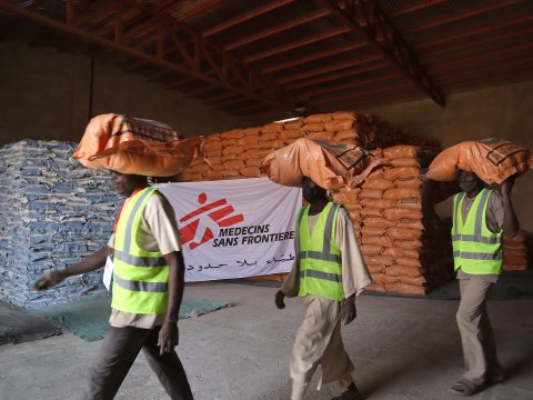 Tre personer bär mat på huvudena i ett lager med en Läkare Utan Gränser-flagga i bakgrunden