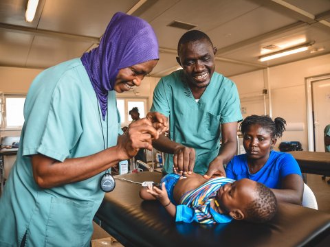 Två personer i kirurggröna kläder undersöker ett barn som ligger på en brits. Bredvid barnet en kvinna som sitter ner vid britsen.