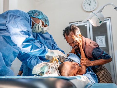Två personer i blå sjukhuskläder och munskydd undersöker ett barn som ligger på en brits. Bredvid barnet en man som håller i barnets arm och lutar sig över barnet.