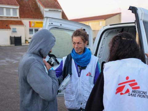 En kvinna i Läkare Utan Gränser-väst står vid en bil med öppen bakdörr och håller handen på en man i grå tröjas axel. 
