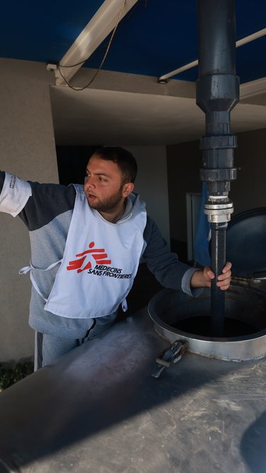 En man i Läkare Utan Gränser-väst står vid en vattentank och pekar.