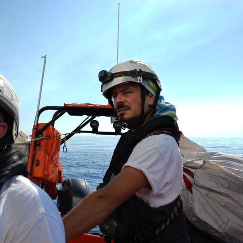 En man med flytväst och hjälm kollar mot kameran medan han står i en båt med öppet hav bakom ryggen